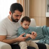 Молодой отец сидит на светлом диване с малышом на коленях и рассматривает красные пятнышки на его предплечье